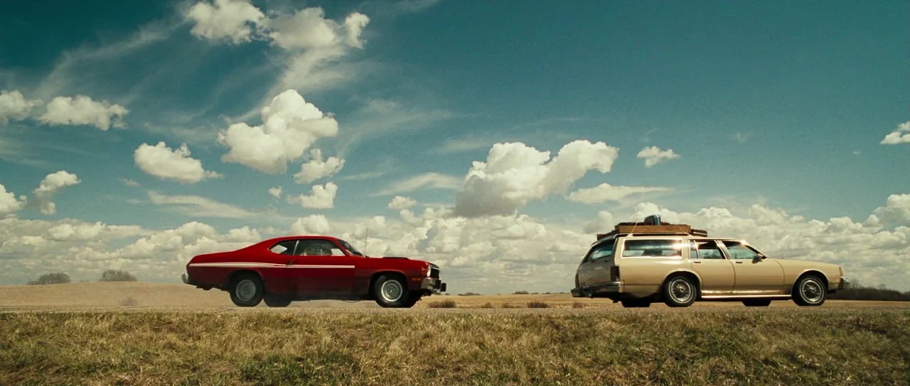 Surveillance (2008) movie still: wide — A desolate paved road cuts through flat, dry fields under a vast cloud-filled sky. A red muscle car drives…