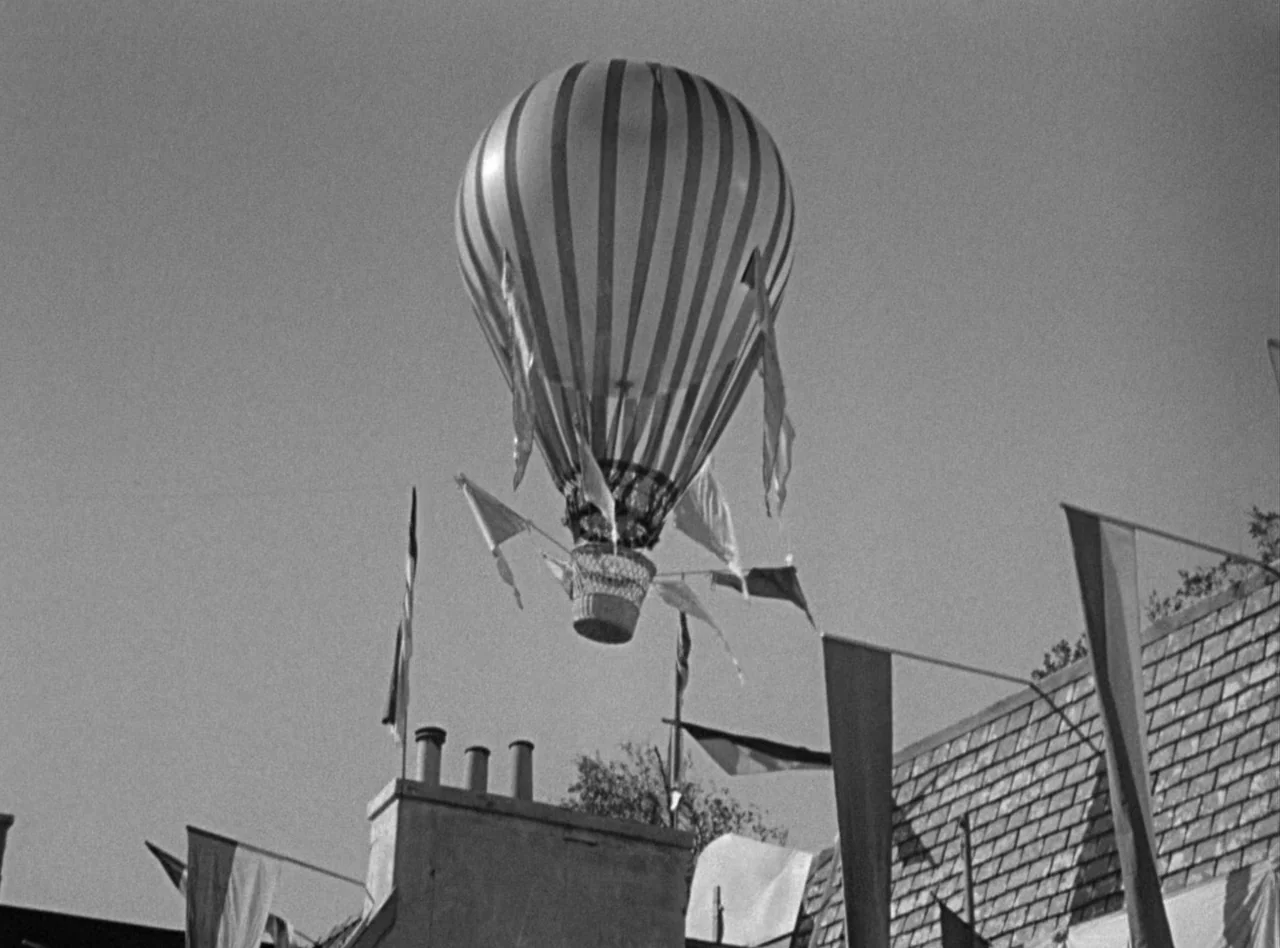 Madame Bovary (1949) movie still: wide — A striped hot air balloon drifts over a house with a tile roof. Numerous flags hang from the building and…