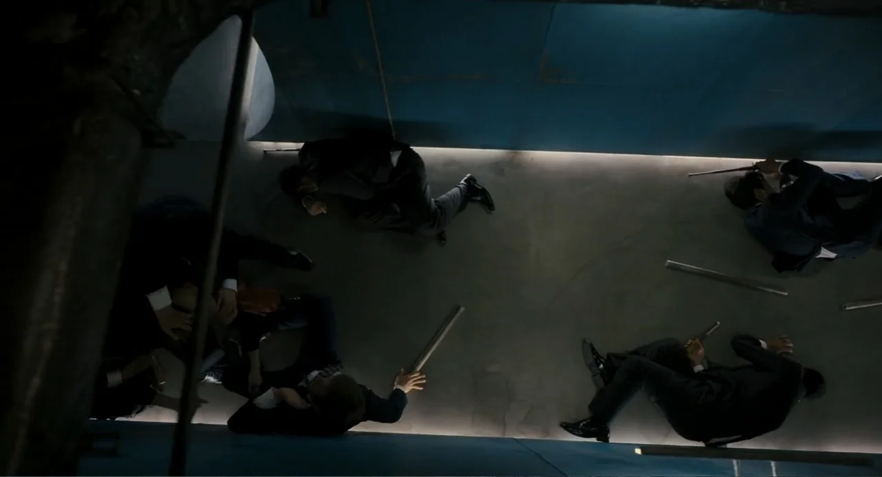 Knuckle Girl (2023) movie still: wide — A narrow, blue-walled corridor features four men lying on the floor in suits. Each figure is positioned…