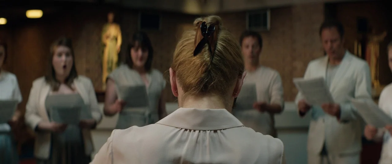 Beast (2017) movie still: medium — A group of people stands in a dim church with wooden paneled walls. A woman seen from behind leads the choir…