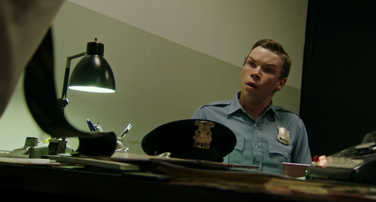 Detroit (2017) movie still: medium — A sparse police station office features pale green walls and a cluttered desk. An officer in a blue uniform…