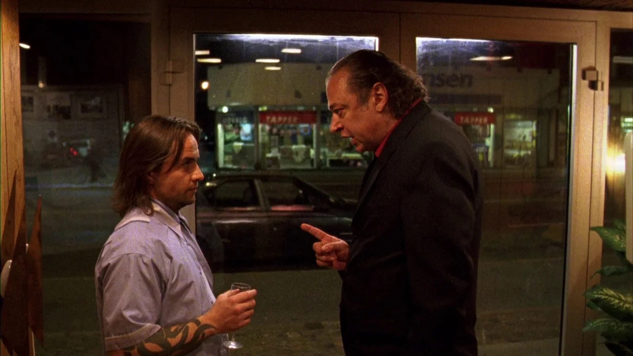 Pusher III (2004) movie still: medium — A man in a blazer confronts a younger man in a shirt near large glass windows. Outside, the night street…