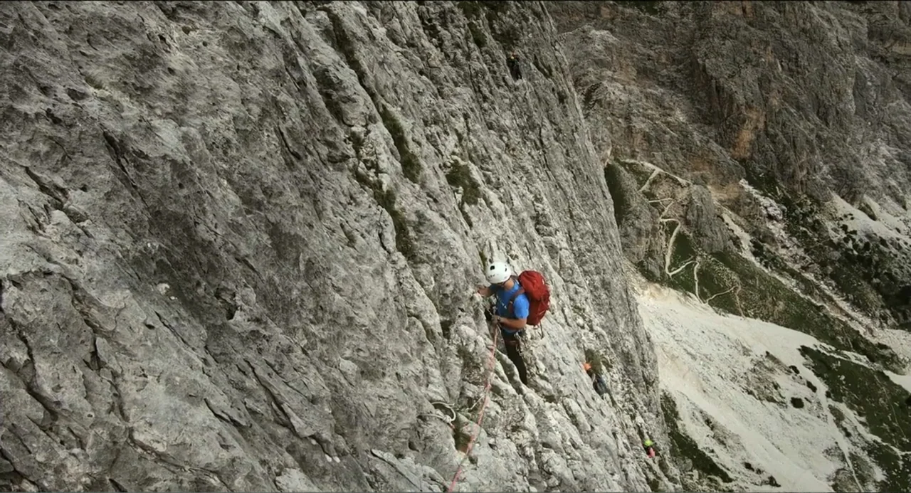 Crooked Trees Gon Give Me Wings (2022) movie still: wide — A sheer mountain rock face rises vertically with a climbing path carved below. A climber…