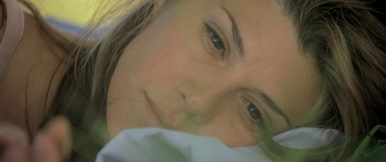 In the Bedroom (2001) movie still: closeup — A woman rests on the ground amidst patches of grass. The frame captures her face from a close angle…