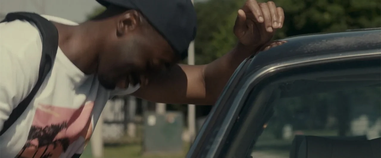 Jump, Darling (2022) movie still: medium — A sunny outdoor parking lot with green trees visible in the soft-focus background. A man leans his weight…