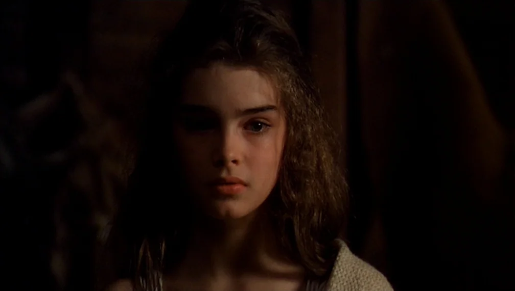 Pretty Baby (1978) movie still: closeup — A dimly lit interior space features a young woman centered in the frame. She stares forward while her long…