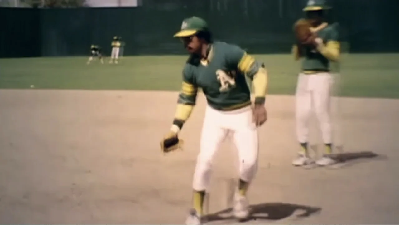 Reggie (2023) movie still: fullbody — A dusty baseball infield features players in green and gold uniforms during practice. One player leans low to…