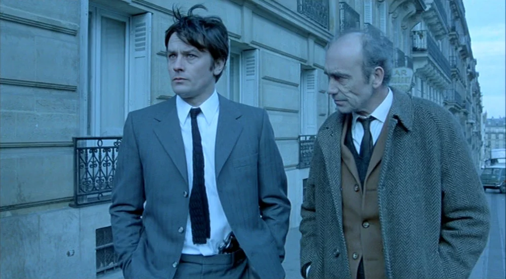 Un Flic (1972) movie still: medium — A Parisian street with classic Haussmann-style stone building facades and iron balcony railings. Two men in…