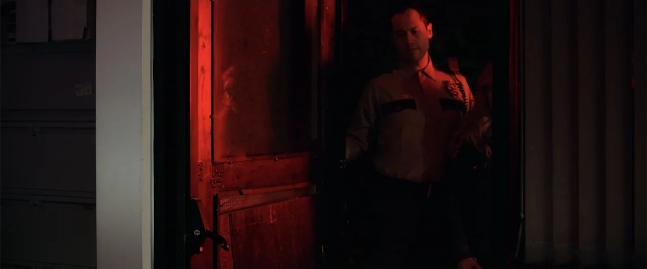 Init!ation (2021) movie still: medium — A dark interior threshold is lit by a vibrant red neon light source. A male police officer in uniform enters…