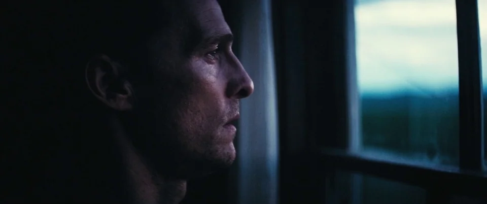 Interstellar (2014) movie still: closeup — An interior space with a dark, indistinct environment framing a large window. A man stands at the glass…