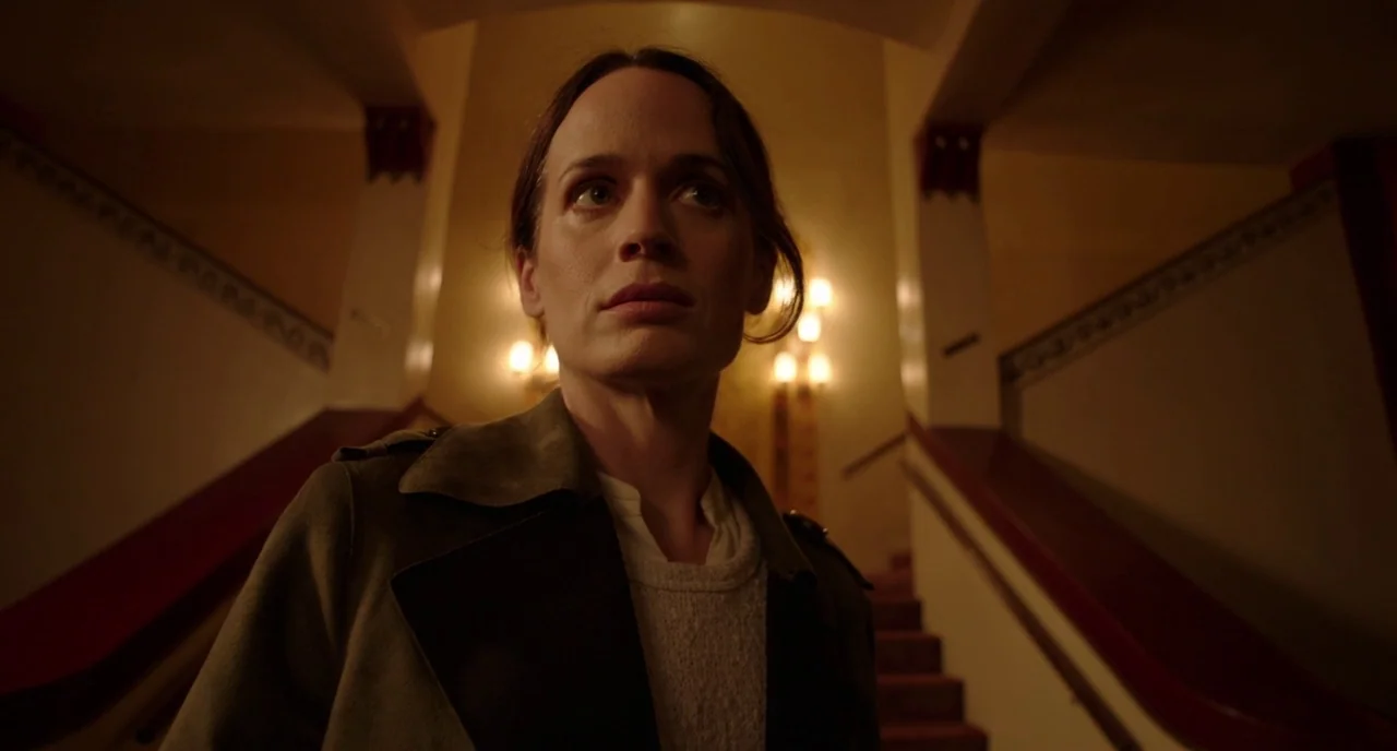 Nightmare Cinema movie still: medium — The interior of a building features a central staircase with wall-mounted lamps. A woman in a coat stands…