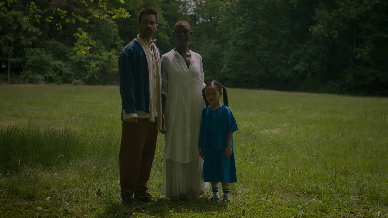 After Yang (2021) movie still: fullbody — A grassy clearing surrounded by a dense forest under soft overcast daylight. Three individuals stand…