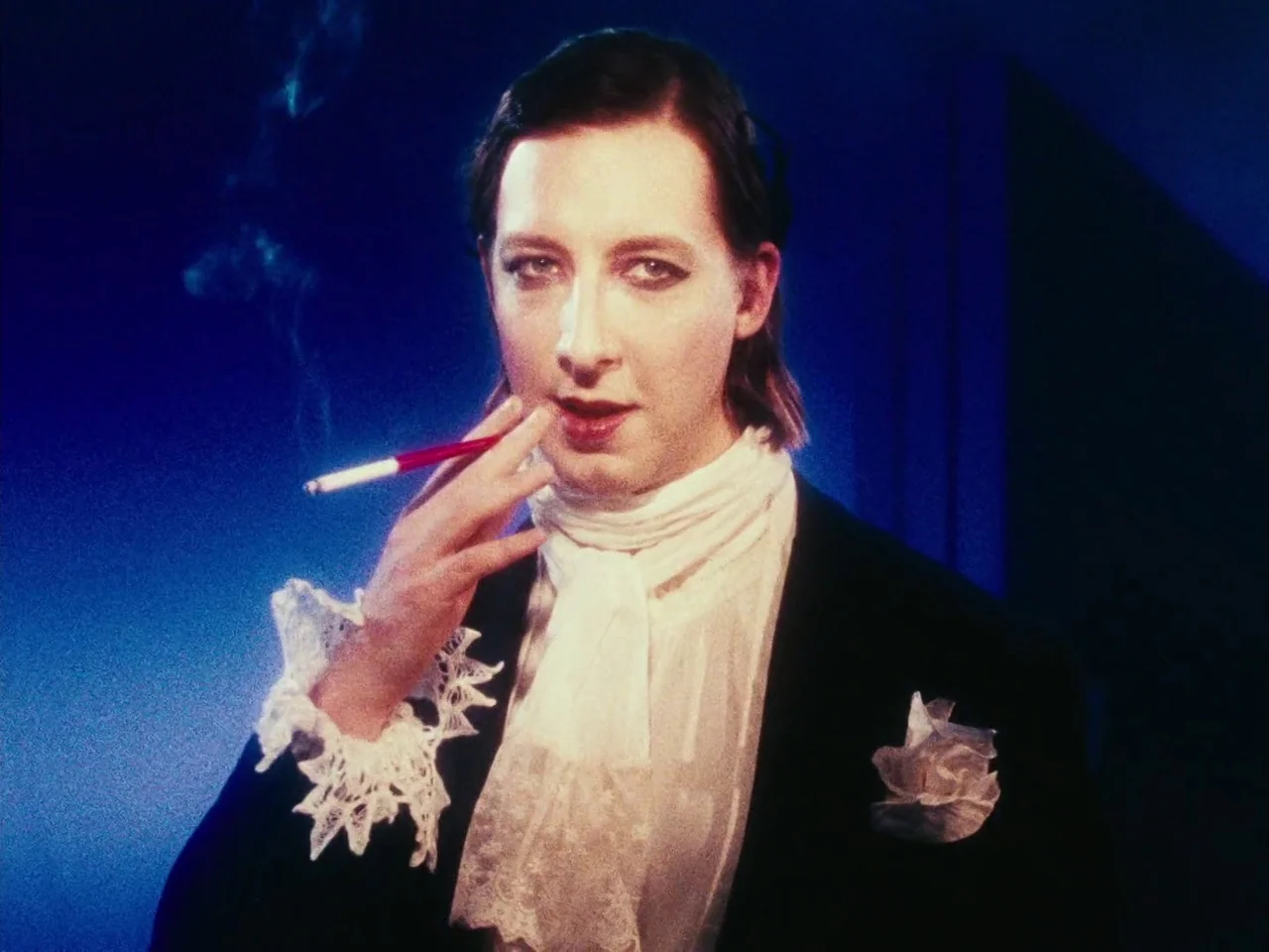 The Twentieth Century (2019) movie still: closeup — A studio space with a vibrant blue gradient backdrop and theatrical lighting. A man dressed in…