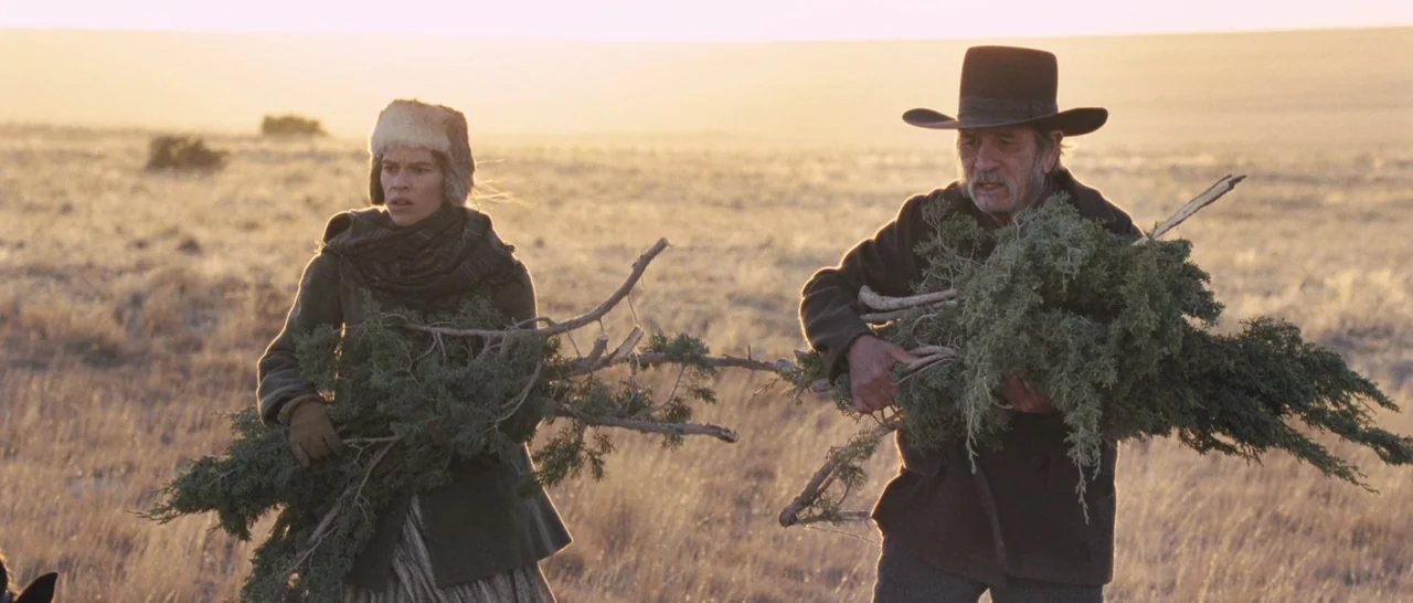 The Homesman (2014) movie still: medium — A wide, sparse field of dry grass glows under the late afternoon sunlight. A woman and an older man carry…