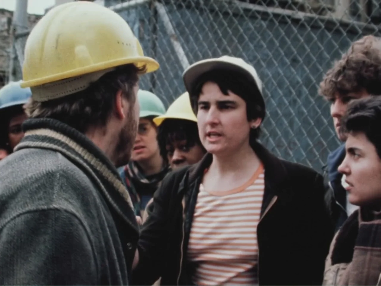 Born in Flames (1983) movie still: medium — A group of workers stands in front of a chain-link fence outdoors. A person with a cap speaks to a man…