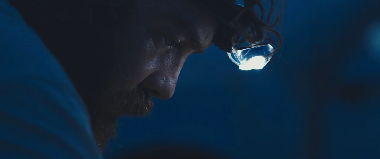 Blue Ruin (2013) movie still: closeup — A dark, dimly lit environment surrounds a man wearing a bright LED headlamp. The man gazes downward intently…