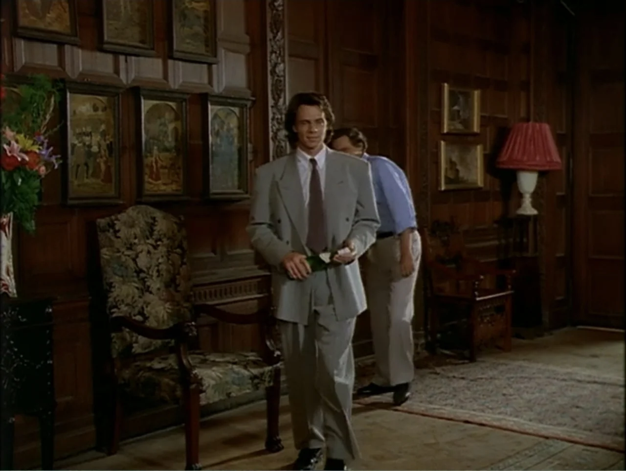 A Change of Place (1994) movie still: fullbody — A wood-paneled hall features ornate framed wall art and a patterned fabric armchair. One man walks…