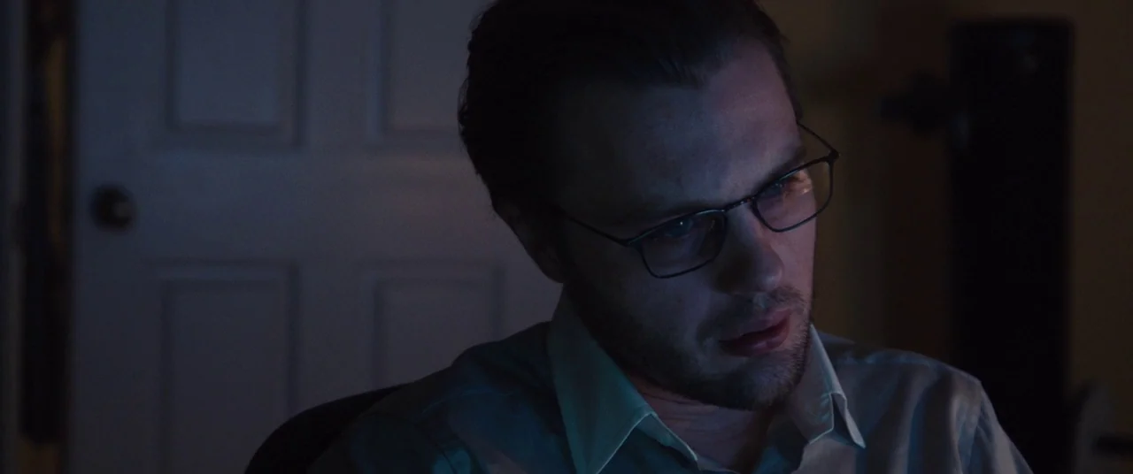 I Origins (2014) movie still: closeup — A dimly lit room at night with a closed white door in the background. A man wearing glasses sits in front of…