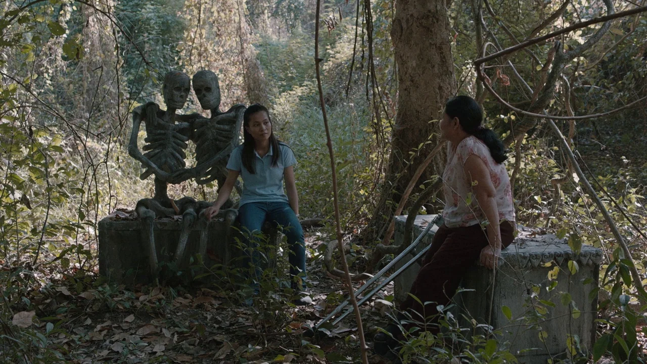 Cemetery of Splendour (2015) movie still: wide — An overgrown, rustic cemetery area features concrete tombs adorned with skeletal statues among…