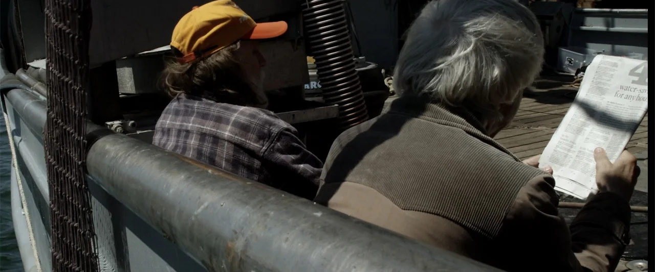 Mermaid Down (2019) movie still: medium — A wooden boat deck under direct sunlight features industrial equipment and metallic piping. Two…