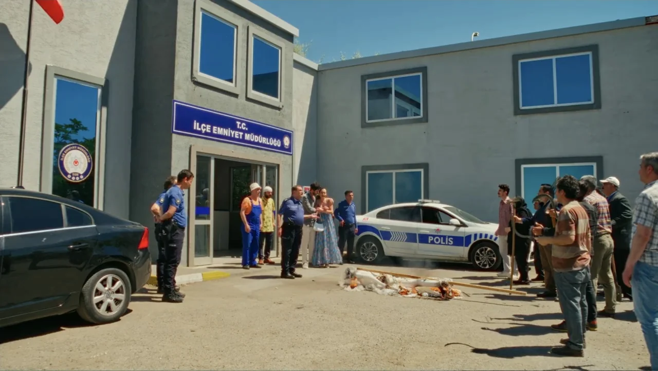 Paranormal Cuma (2024) movie still: wide — A group of people stands outside a gray government building with a blue sign. A small fire burns on the…