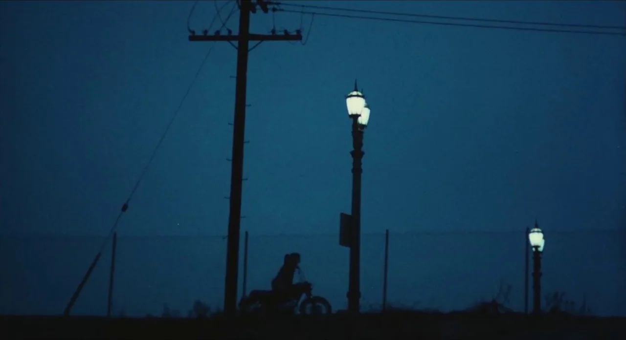 The Other Side of the Wind (2018) movie still: wide — A dark, empty roadside at night features a lone motorcyclist riding past streetlamps. Power…