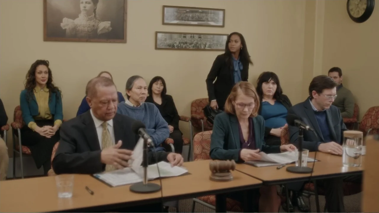 A Christmas Break (2020) movie still: medium — A formal hearing room features tan walls adorned with framed pictures and a clock. A group of…