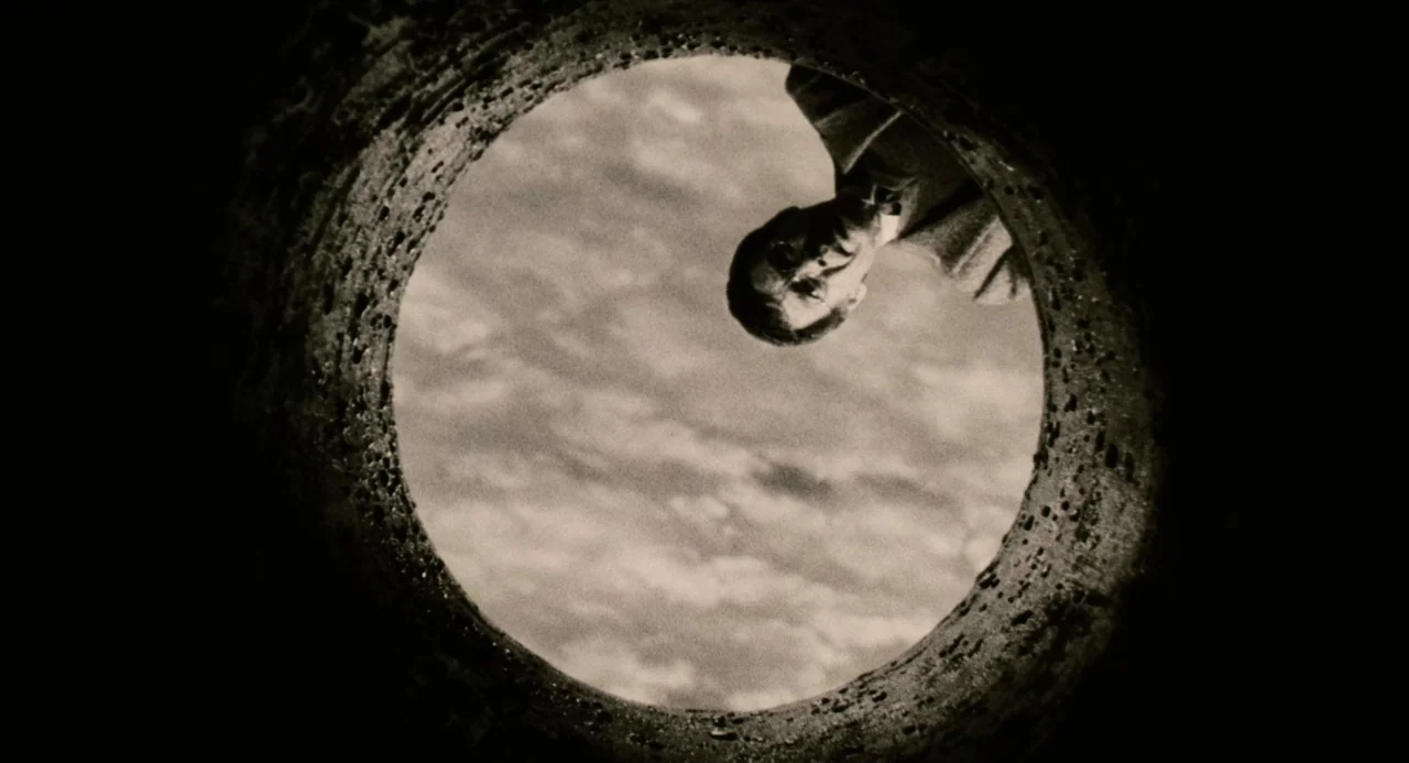 Ringu (1998) movie still: wide — A man peers down from above into the dark, circular frame of a stone well. The sky with light clouds is visible…