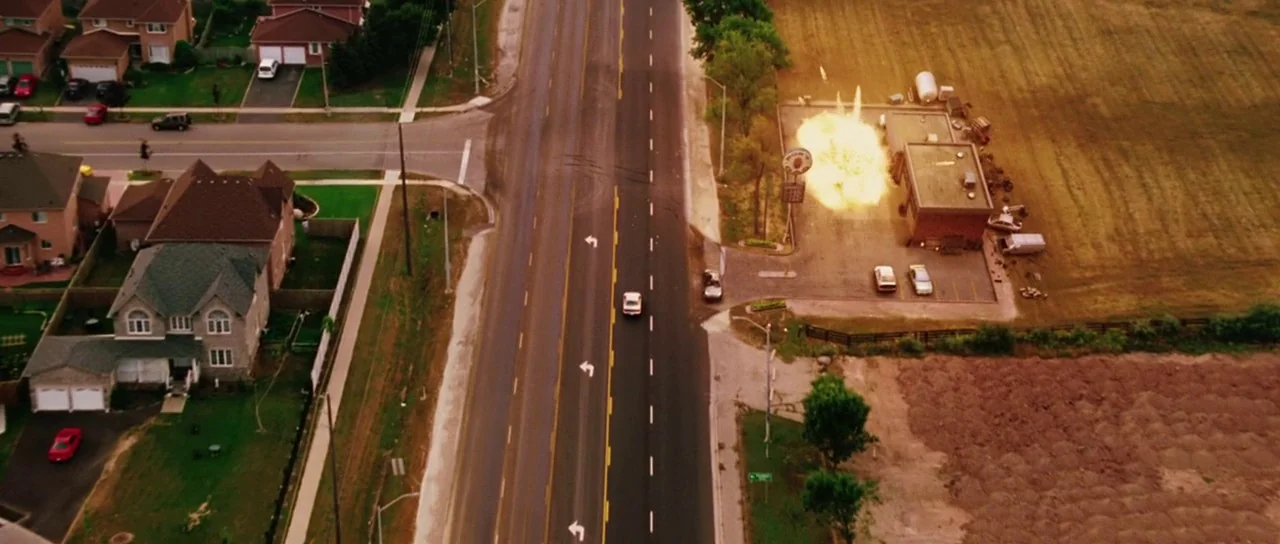 Dawn of the Dead (2004) movie still: establishing — A top-down view shows a suburban landscape with a road bordering a large field. An explosion…