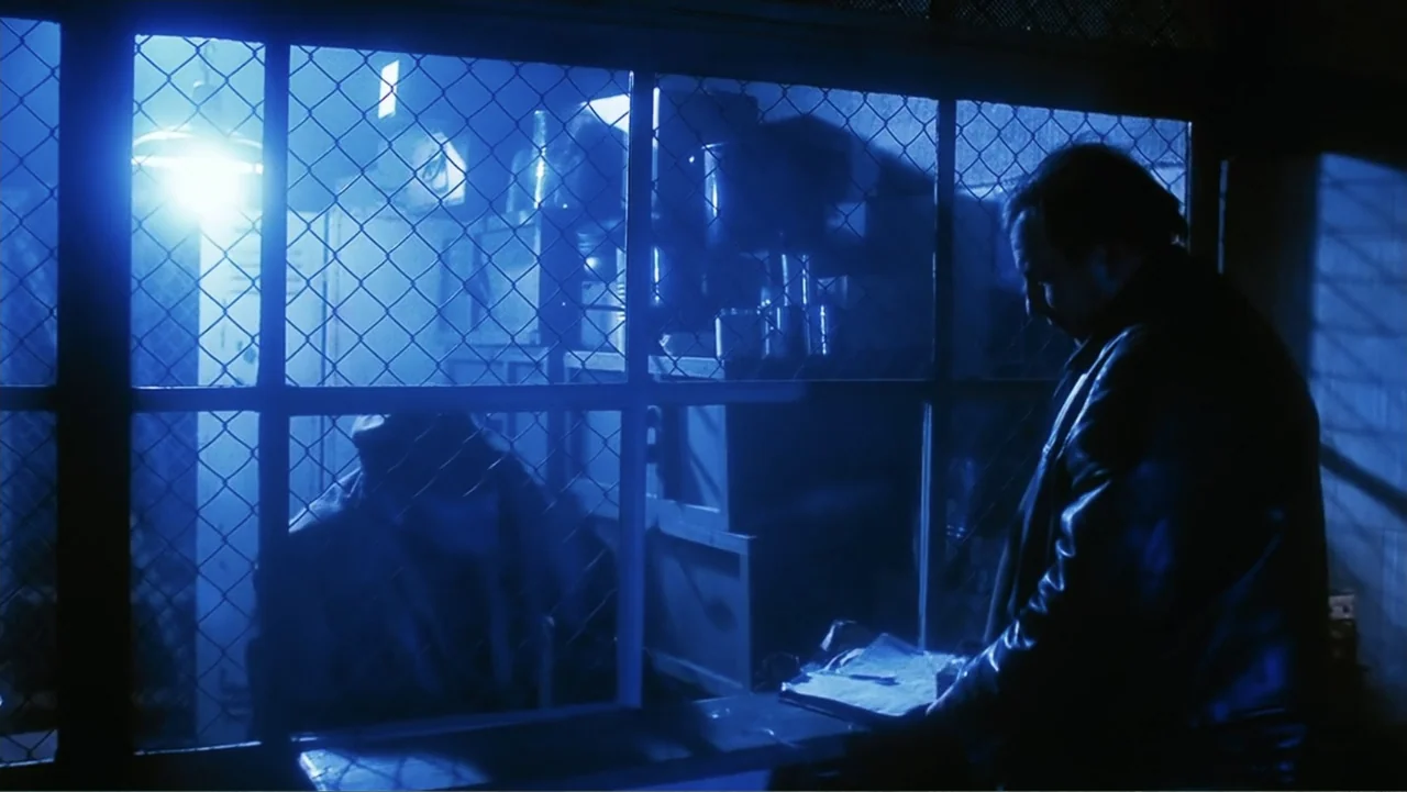 O-Bi, O-Ba: The End of Civilization (1985) movie still: medium — A stark indoor setting features a wire mesh partition separating a service area.…