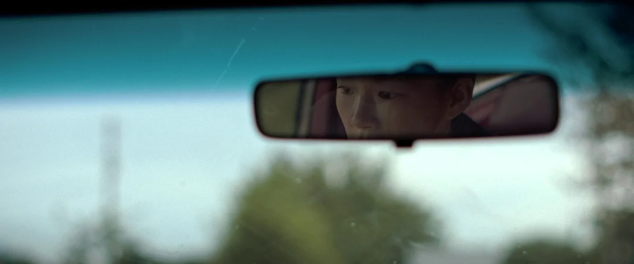 Minari (2020) movie still: closeup — The interior of a moving vehicle shows the driver reflected in the rearview mirror. The reflection is centered…