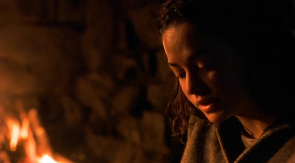 Limbo (1999) movie still: closeup — A dark stone interior lit only by the glowing light of a nearby fire. A woman sits in front of the flames while…