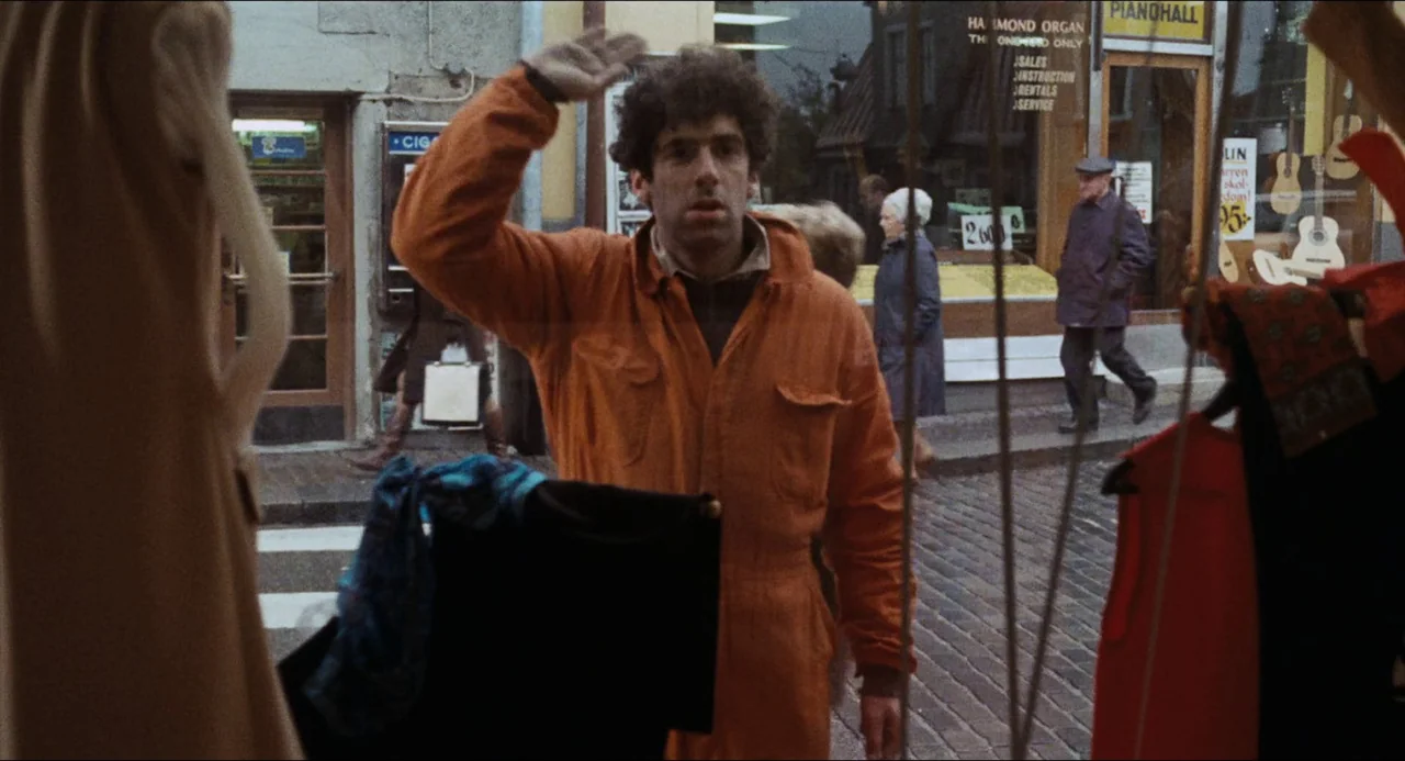 The Touch (1971) movie still: medium — A man in an orange jumpsuit walks past a storefront window on a city street. Passersby are visible in the…