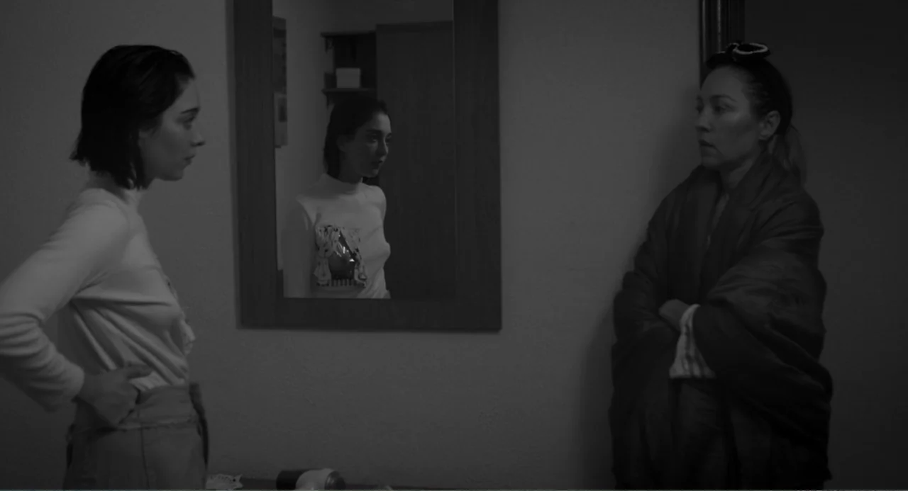 El Planeta (2021) movie still: medium — A dimly lit room contains a mirror hanging on a plain wall. Two women stand opposite each other while one…