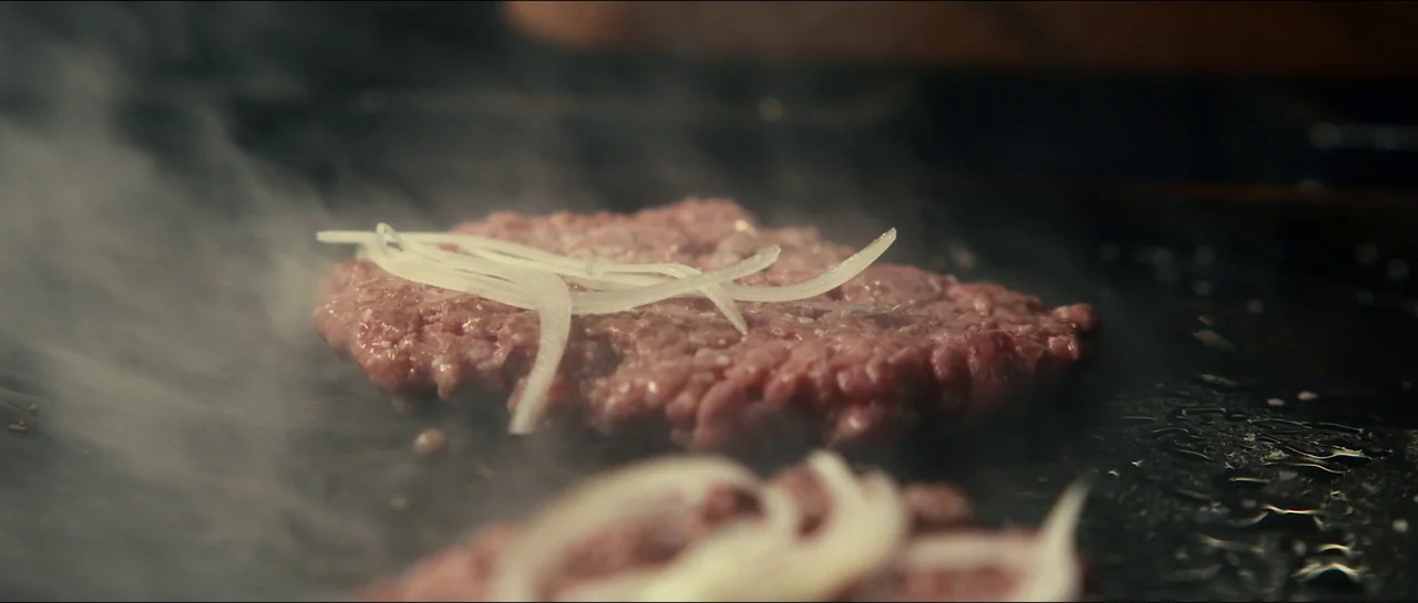 The Menu (2022) movie still: closeup — A raw ground meat patty sits on a dark, hot metal griddle surface. Thin slices of white onion rest on top of…