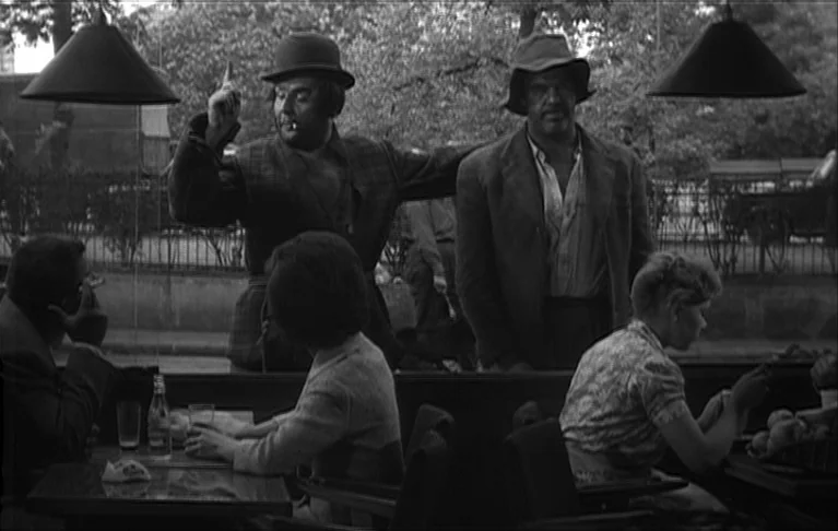 Le Signe du Lion (1962) movie still: medium — An outdoor cafe terrace with two men standing outside a large glass window pane. They gesture toward…