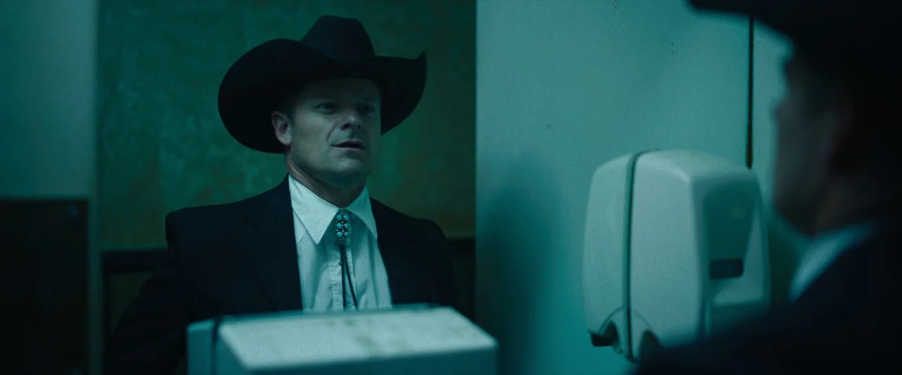 LaRoy, Texas (2023) movie still: medium — A dimly lit, narrow restroom space features cool green-tinted walls and white tile surfaces. A man in a…