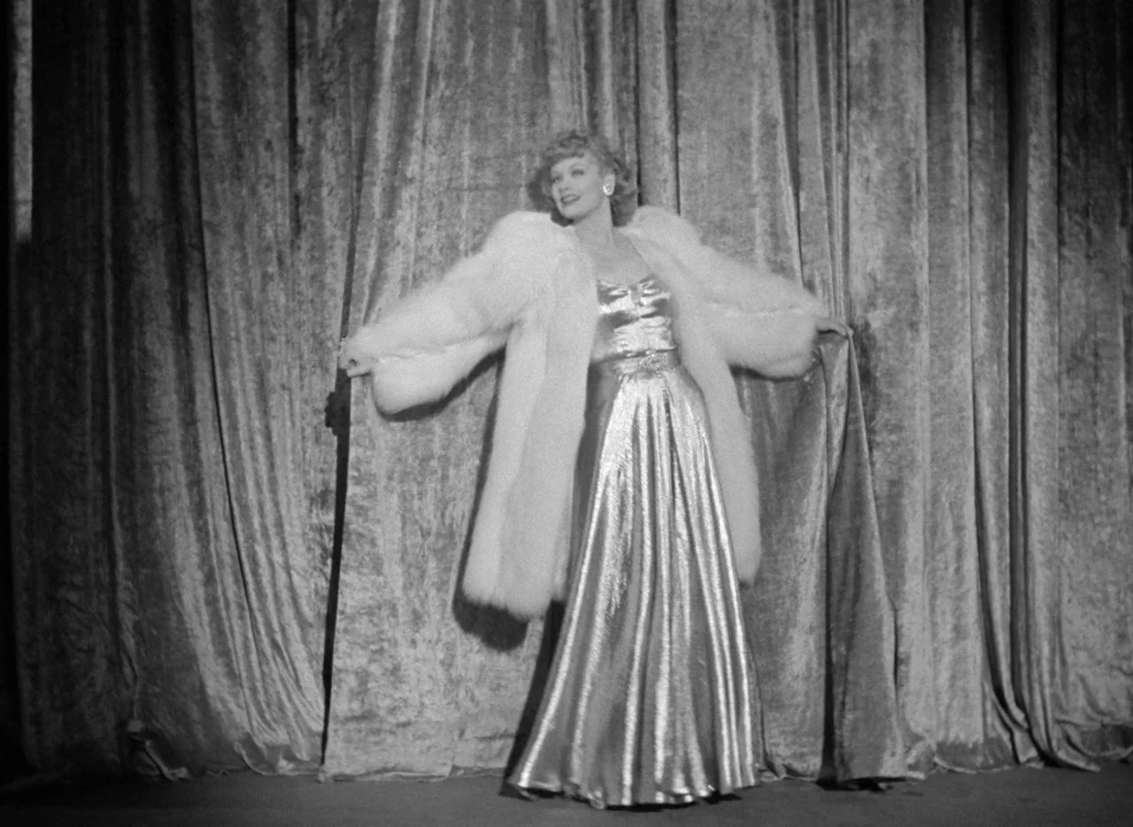 Dance, Girl, Dance (1940) movie still: fullbody — A woman stands centered in front of draped heavy velvet curtains. She holds open a large white fur…