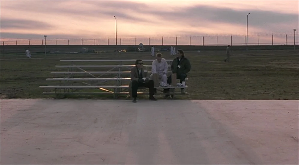 Bottle Rocket (1996) movie still: wide — A flat, open field at sunset is enclosed by a long metal security fence. Three men sit on metal bleachers…