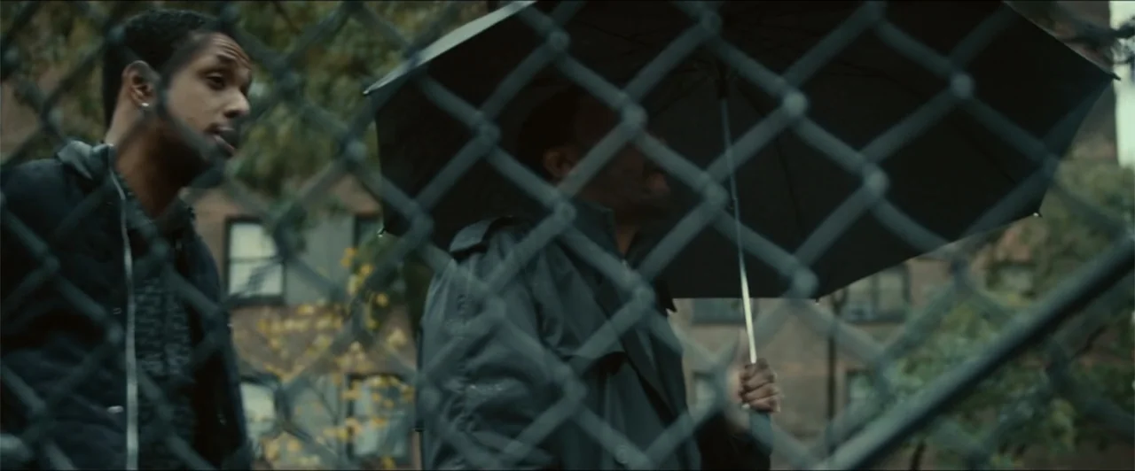 Exposed (2016) movie still: medium — Two men stand outside near an urban chain link fence during an overcast day. One man faces the camera while the…