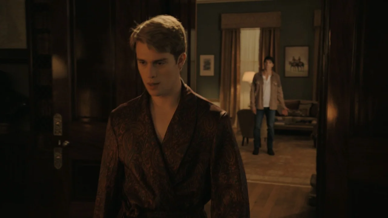 Red, White & Royal Blue (2023) movie still: medium — A dark, wood-paneled residential hallway connects to a warmly lit living area in back. A man in…