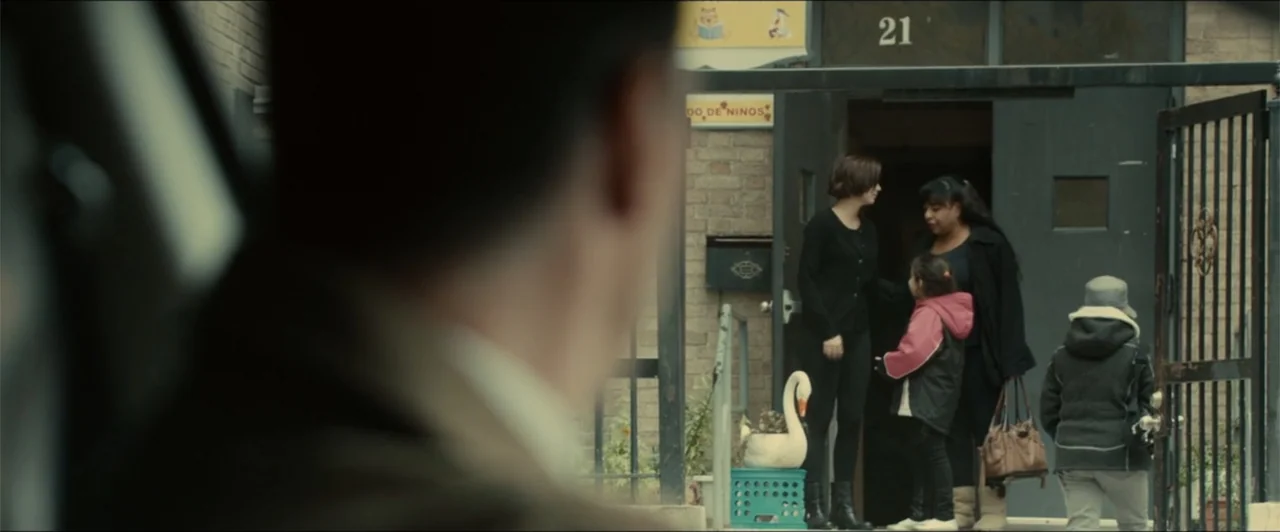 Exposed (2016) movie still: medium — A brick building entrance with a black metal gate and hanging address number 21. Three people stand together at…