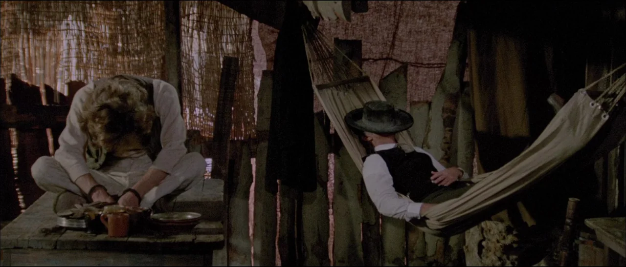 The Grand Duel (1972) movie still: medium — A rustic shelter with walls constructed of woven reeds and vertical wooden planks. One man sits hunched…