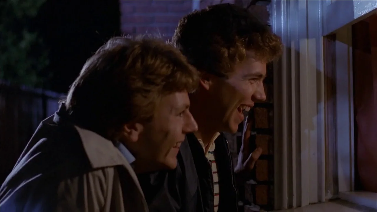 The Slumber Party Massacre (1982) movie still: medium — Two young men stand outside a residential house at night peering through a window. The men…