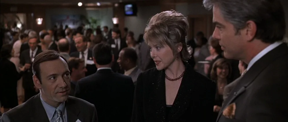 American Beauty (1999) movie still: medium — A crowded social gathering occurs in a dimly lit, large carpeted function room. Three people stand in…