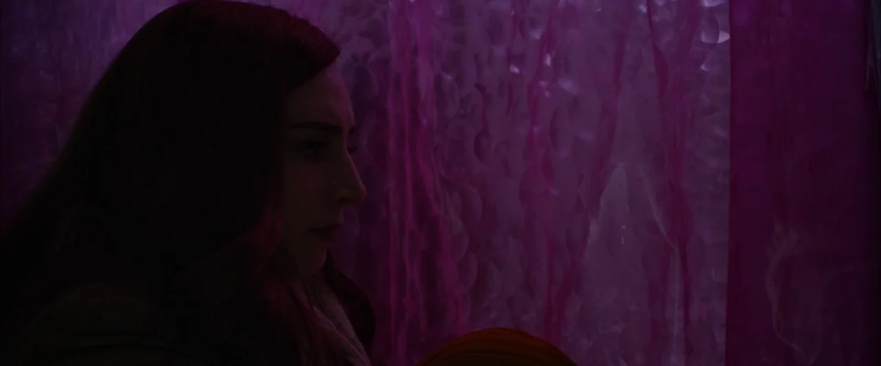 The Stylist (2020) movie still: closeup — A woman stands in a dark space beside a textured, translucent magenta surface. She gazes to the right…