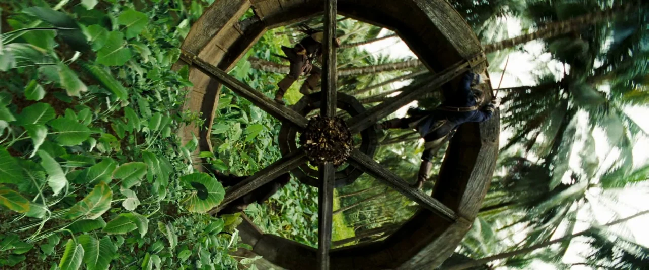 Pirates of the Caribbean: Dead Man’s Chest movie still: wide — A large wooden water wheel rotates vertically through dense tropical jungle foliage.…