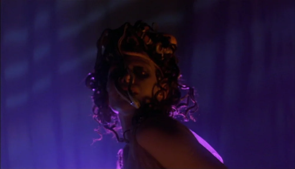 Bram Stoker’s Dracula (1992) movie still: closeup — A woman stands in a dimly lit, purple-hued space with abstract lighting patterns. Several snakes…