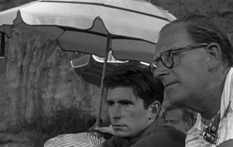 On The Beach (1959) movie still: medium — Two men sit together under a beach umbrella near a rocky cliffside. The older man with glasses looks away…