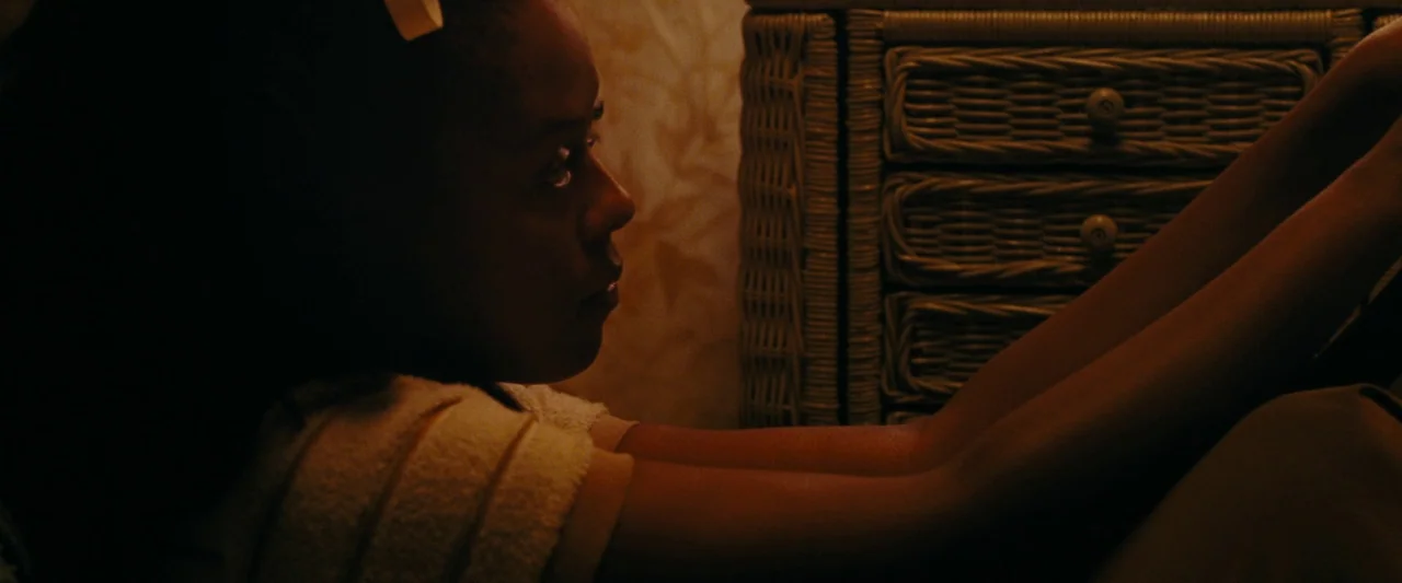 All Dirt Roads Taste of Salt (2023) movie still: medium — A dimly lit space features a wicker chest of drawers with floral-patterned wallpaper…